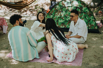 Four friends are gathered on a picnic blanket near a tent and enjoying sunny weather. The setting...