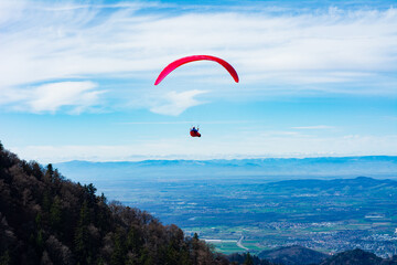 Soaring high above the valley, a bright red paraglider embraces the freedom of flight against a backdrop of stunning mountains and endless sky