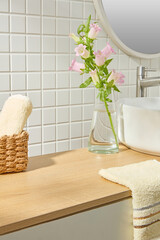 Close-up of natural-toned bathroom with basket, towel layers, white sink and flowers in glass vase....