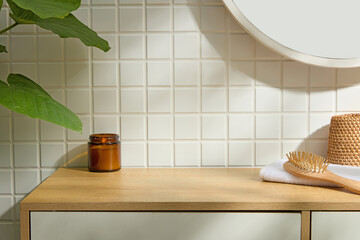 Wood counter with amber glass jar and towel under round mirror. White mosaic tiles and leafy plant...