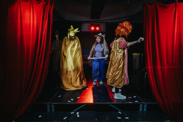 Actors dressed in vibrant costumes perform on a stage framed with red curtains, creating a lively atmosphere and showcasing playful expressions during a theatrical performance.