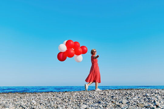 Blond woman in red polka dot dress holding balloons on a pebble beach by the Ionian Sea on a sunny day.