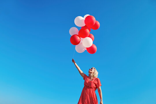 Blond woman in a red polka dot dress holding red and white balloons under a clear sky on a sunny day