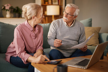 In the daytime, a senior couple sits on a sofa in their living room. The man reviews documents...