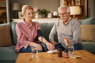 An elderly couple sits comfortably on a sofa, with the woman using a blood pressure monitor, while the man explains the process, surrounded by health supplements and glasses of water.