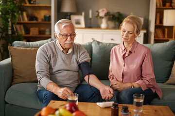 An elderly man measures his blood pressure while sitting on the sofa in his living room. His wife sits next to him while he is taking the measurement with the digital monitor.