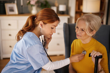 A home care nurse in scrubs uses a stethoscope to listen to the heart and lungs of an elderly woman...
