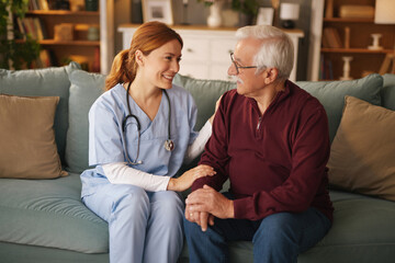 A female home health Nurse sits beside a senior man on a couch in a living room. The Nurse smiles...