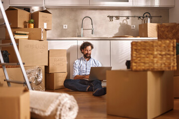 An individual sits on the floor of a modern kitchen, using a laptop while surrounded by cardboard...