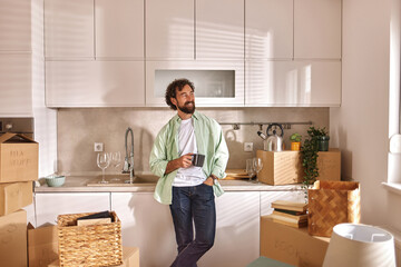 A man relaxes in a bright kitchen filled with moving boxes and unpacked items. He holds a coffee...