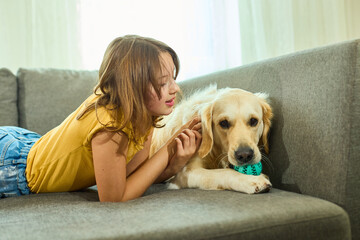 A cheerful teen girl relaxes on a sofa beside her golden retriever, who is happily chewing a ball....