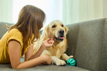 A cheerful teen girl relaxes on a sofa beside her golden retriever, who is happily chewing a ball....