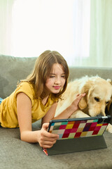 A teen girl lies comfortably on the couch in a cozy living room, engaging with her tablet. Her...