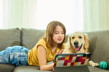 A teen girl lies comfortably on the couch in a cozy living room, engaging with her tablet. Her...
