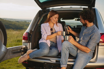 A couple sits comfortably in the trunk of their car, sharing drinks and smiling at each other,...
