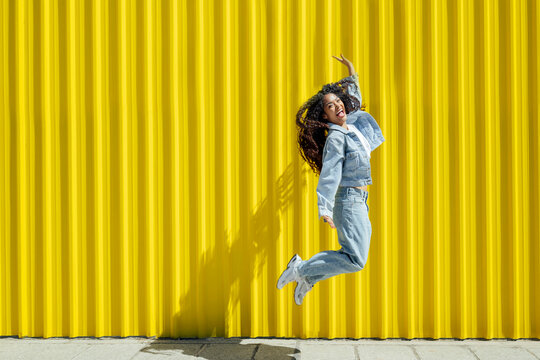 Woman in denim jumping joyfully against a yellow urban background