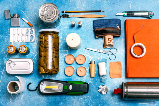 Flat lay of survival essentials kit with candles, lighter, batteries, canned food, and solar flashlight on a blue background