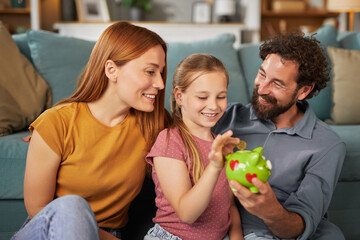 A family sits together on a couch, sharing smiles and laughter as they help a young girl count coins in a colorful piggy bank during a cozy evening at home.