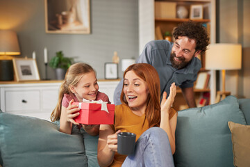 A cheerful family shares a joyful moment on a couch as a girl presents a red gift to her surprised...