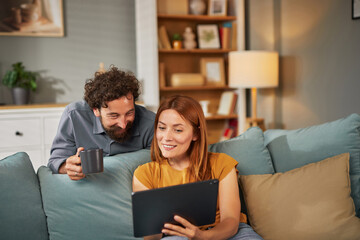 Two people sit comfortably on a soft couch, sharing laughter as they engage with a tablet, creating...