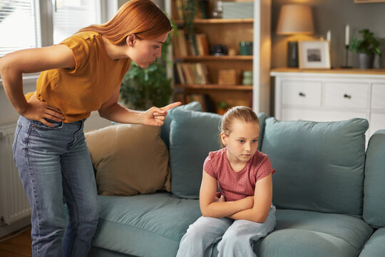 A mother is reprimanding her young daughter in a comfortable living room setting. The girl sits with crossed arms, visibly upset, as her mother expresses disappointment.