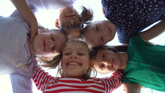Low angle view of happy little children in circle look at camera embrace and laugh in summer park. Team kids play outdoors. Close up of face girls and boys. Schoolchildren on holiday