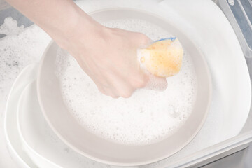 Female hand with sponge for washing dishes and bowls in water with foam or dishwashing liquid. washing dishes in the sink