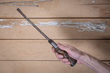 Old flathead screwdriver with wooden handle held by hand over rustic beige wooden table. The worn...