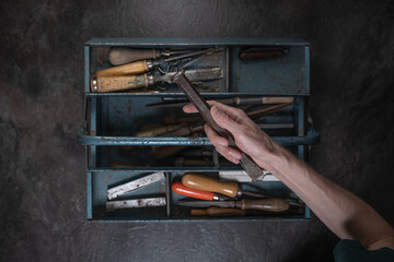 A hand holds a vintage iron chisel above an open metal toolbox filled with old tools, photographed...