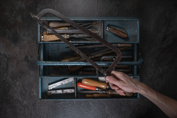 A hand holds a vintage bow saw above an open metal toolbox filled with old tools, photographed from...
