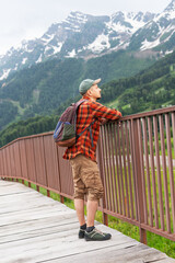 Young man in plaid shirt , backpack and cap leans against a railing, gazing at a breathtaking mountain landscape, showcasing nature's beauty and adventure spirit