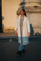 Woman in stylish coat walking confidently near grand architectural building during sunset, exuding...