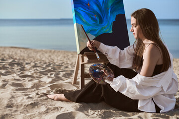 Young female artist paints on beach, capturing ocean inspiration on canvas. Peaceful ambiance...