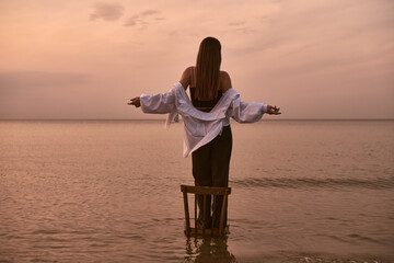 Young woman stands on stool in shallow sea, arms wide, embracing tranquil sunset. Warm hues and...