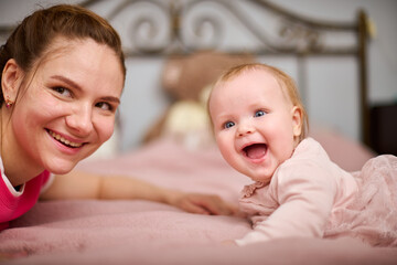 Young woman with brown hair and smiling baby girl on pink bed. Light-hearted interaction, creating joyful atmosphere. Warm lighting enhances bond