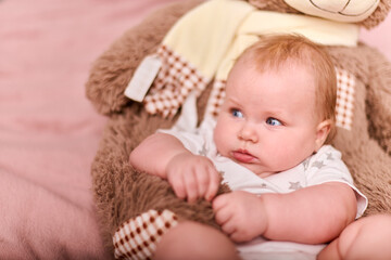 Baby girl, light hair, caucasian, lying on pink blanket, cuddling toy bear. Soft lighting adds...
