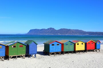 Brightly painted beach huts at Muizenberg Beach, South Africa, with ocean waves and mountains in...