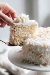 Close-up of a hand serving a moist slice of coconut cake with creamy frosting and toasted coconut flakes, captured in soft natural light for a fresh, homemade feel.
