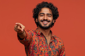 A smiling man with curly hair and a beard, wearing a colorful patterned shirt, points directly at the camera against a solid orange background.