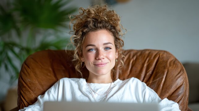 A joyful woman with curly hair smiles warmly while sitting in a large cozy armchair, emanating a sense of comfort and contentment in her relaxed living space.