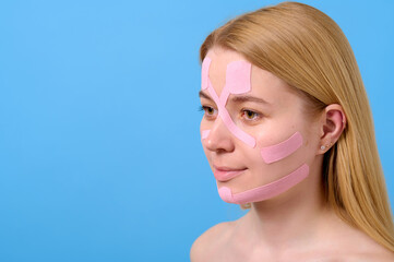 woman with pink facial tape demonstrates skincare technique against blue background, highlighting...
