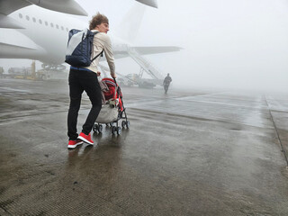 Man is pushing a stroller on a wet runway. The stroller is red and has a yellow stripe near...