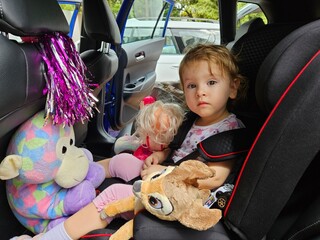 Young girl is sitting in a car seat with a stuffed animal. There are two other stuffed animals in the car seat safety chair with belt