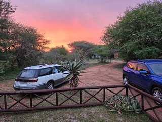 Blue car is parked in front of a wooden fence, with a white car parked behind it. The scene takes...