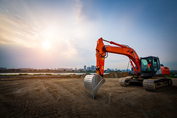 Orange excavator parked on construction site with city skyline and sunset in background
