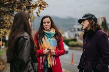 Three women are chatting outdoors in a city setting, dressed stylishly in winter clothing,...