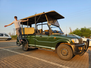 Man boy is standing on the tire roof of a green vehicle. The vehicle is parked in a parking lot...