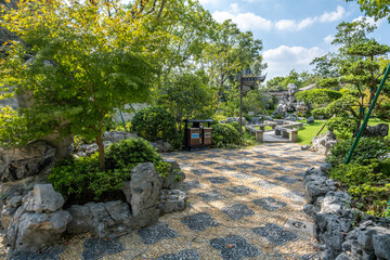 Scenic Chinese garden in a public park, featuring traditional stone landscaping, patterned mosaic...