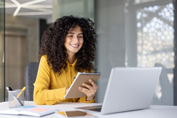A smiling woman with curly hair takes notes while working on a laptop in a modern office setting.