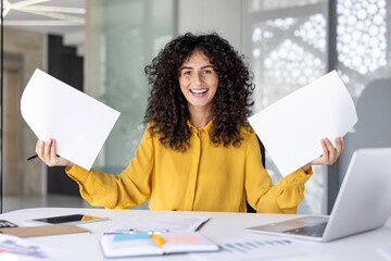 A joyful woman in a yellow shirt holds up blank papers, celebrating with a big smile in an office...